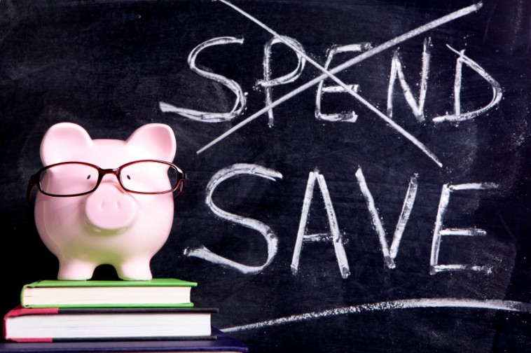 Pink piggy bank with glasses standing on books next to a blackboard with simple spend and save message. Sharp focus on the piggy bank with blackboard message slightly blurred.